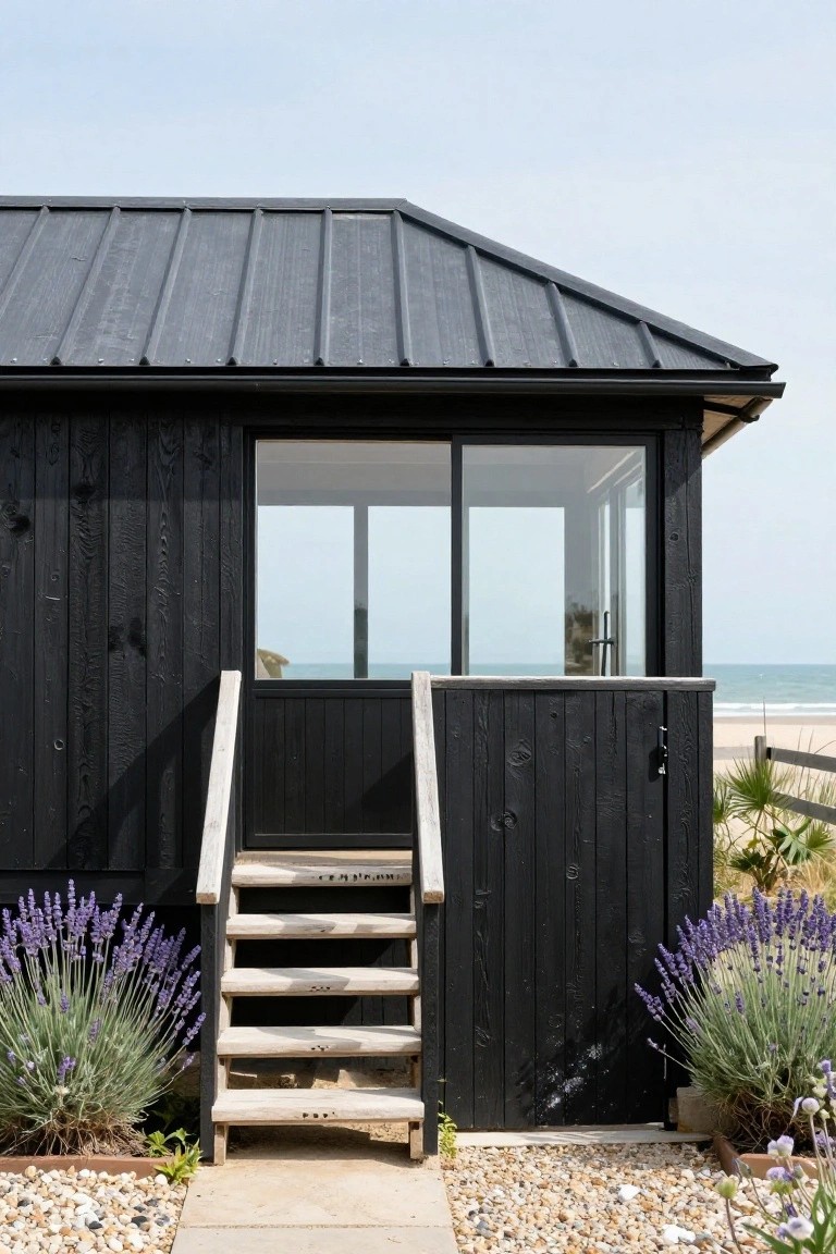Corner view of a black wooden cabin with dark shingle roof, large glass windows facing the ocean, light wood entry steps, lavender plants, and a gravel path.