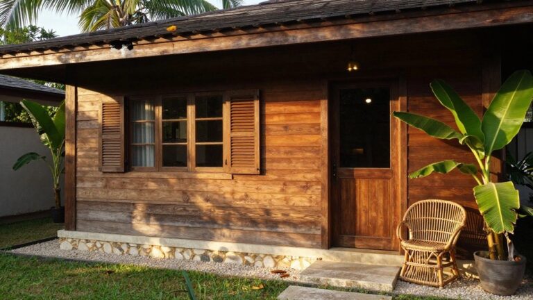 19 Beautiful Tropical Wooden House Design Ideas for Breezy Nature Living Wooden tropical house exterior with slatted side walls, dark paneled door, rattan bench on stone porch steps, gravel ground cover, banana plants, and palm trees nearby.