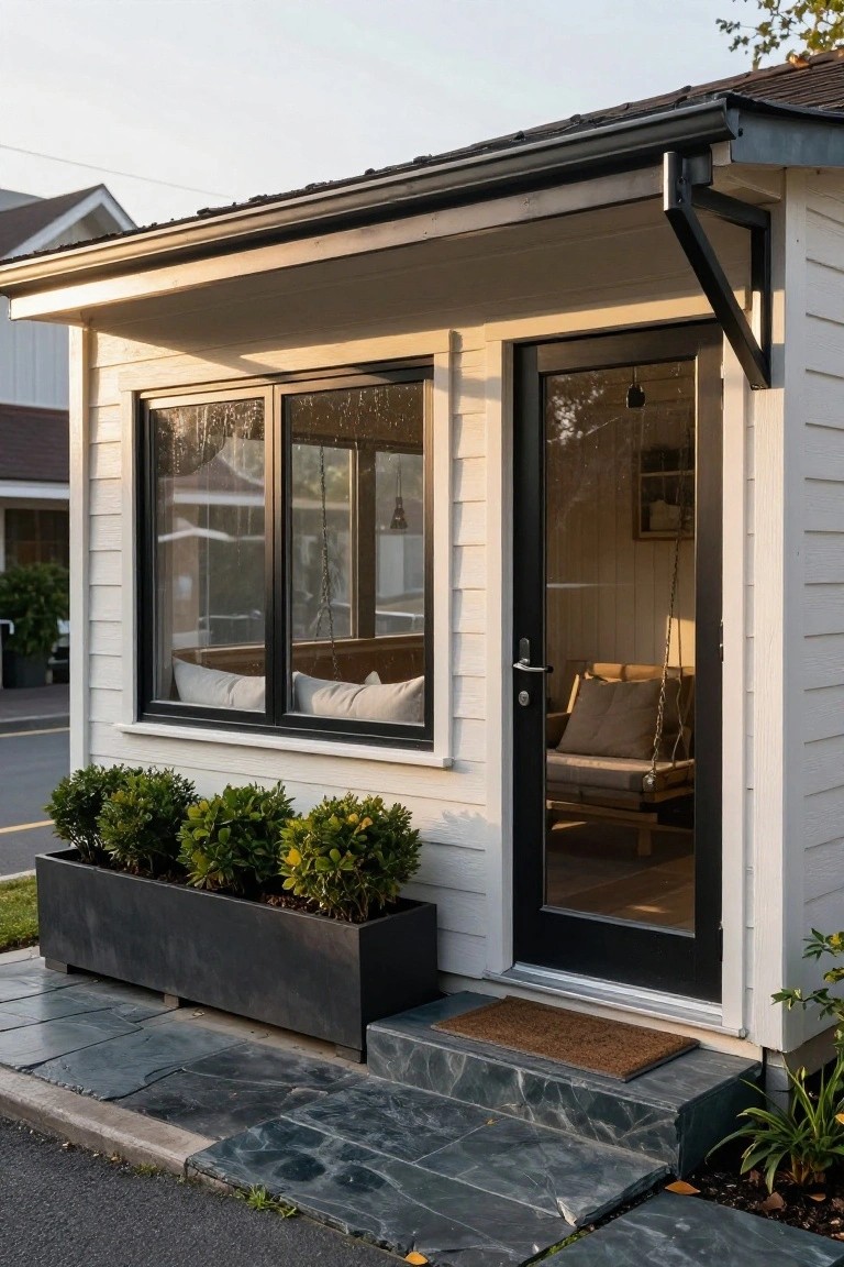 Small white wooden house exterior with black-framed large windows and glass front door under an overhanging roof, boxwood shrubs in a long black rectangular planter beside slate steps leading to a doormat at the entry.