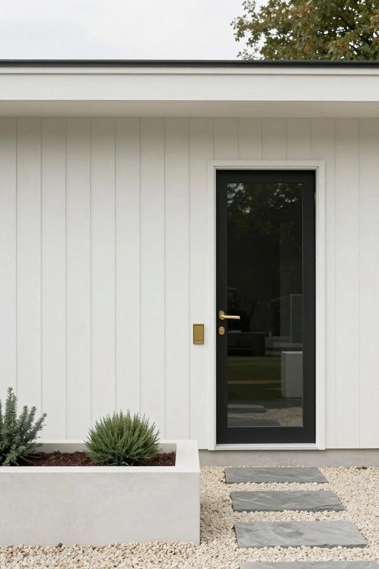 White vertical board-and-batten siding wall with a tall black-framed glass entry door, gold door knob and escutcheon, concrete planter box containing two small green shrubs, gray stone steps on white pebble gravel ground.