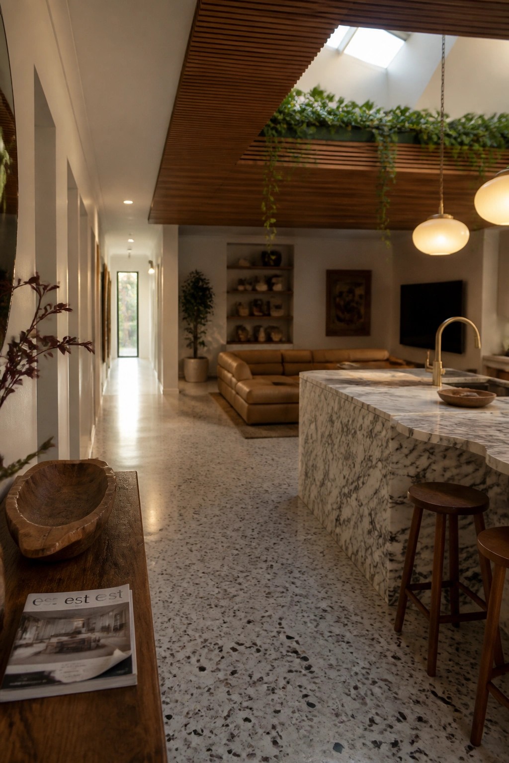 An interior view of a modern kitchen and living space with a wood slat ceiling, hanging plants, marble island, and leather sofa.