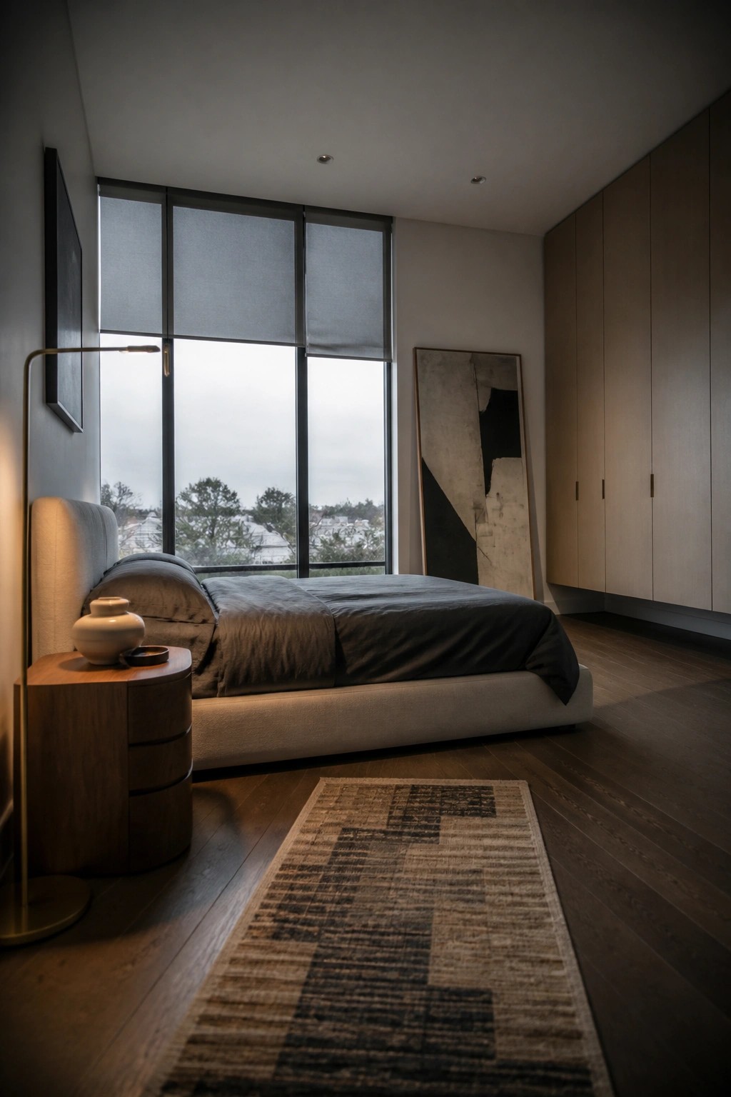 A modern bedroom with dark wood flooring, a large bed with gray bedding, a wooden nightstand, tall light wood cabinets, and a patterned rug on the floor.