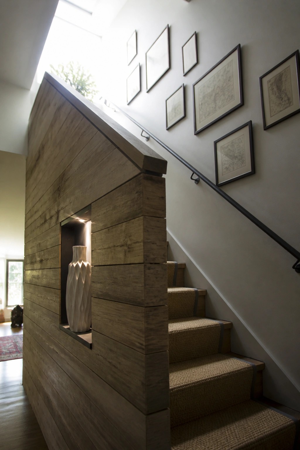 A staircase next to a wood plank wall with a recessed niche holding a white vase and framed pictures on the wall above the steps.