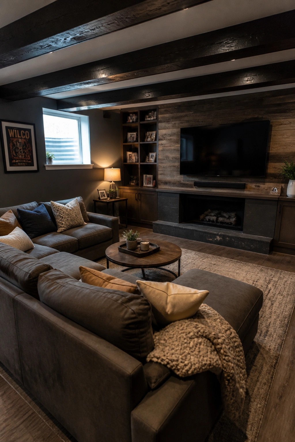 A living room interior with dark faux wood beams on the ceiling, a large gray sectional sofa, round coffee table, built-in shelving, and a fireplace with a television mounted above.