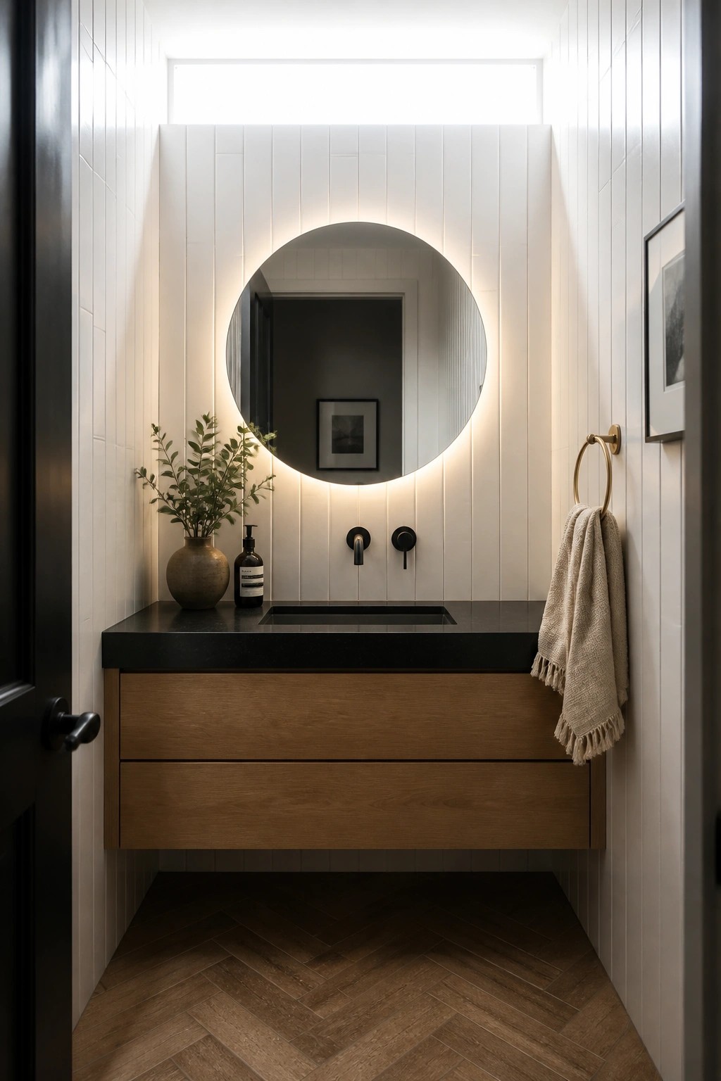 A narrow bathroom featuring dark herringbone wood flooring, a floating wood vanity with black countertop, white vertical shiplap walls, and a round backlit mirror.