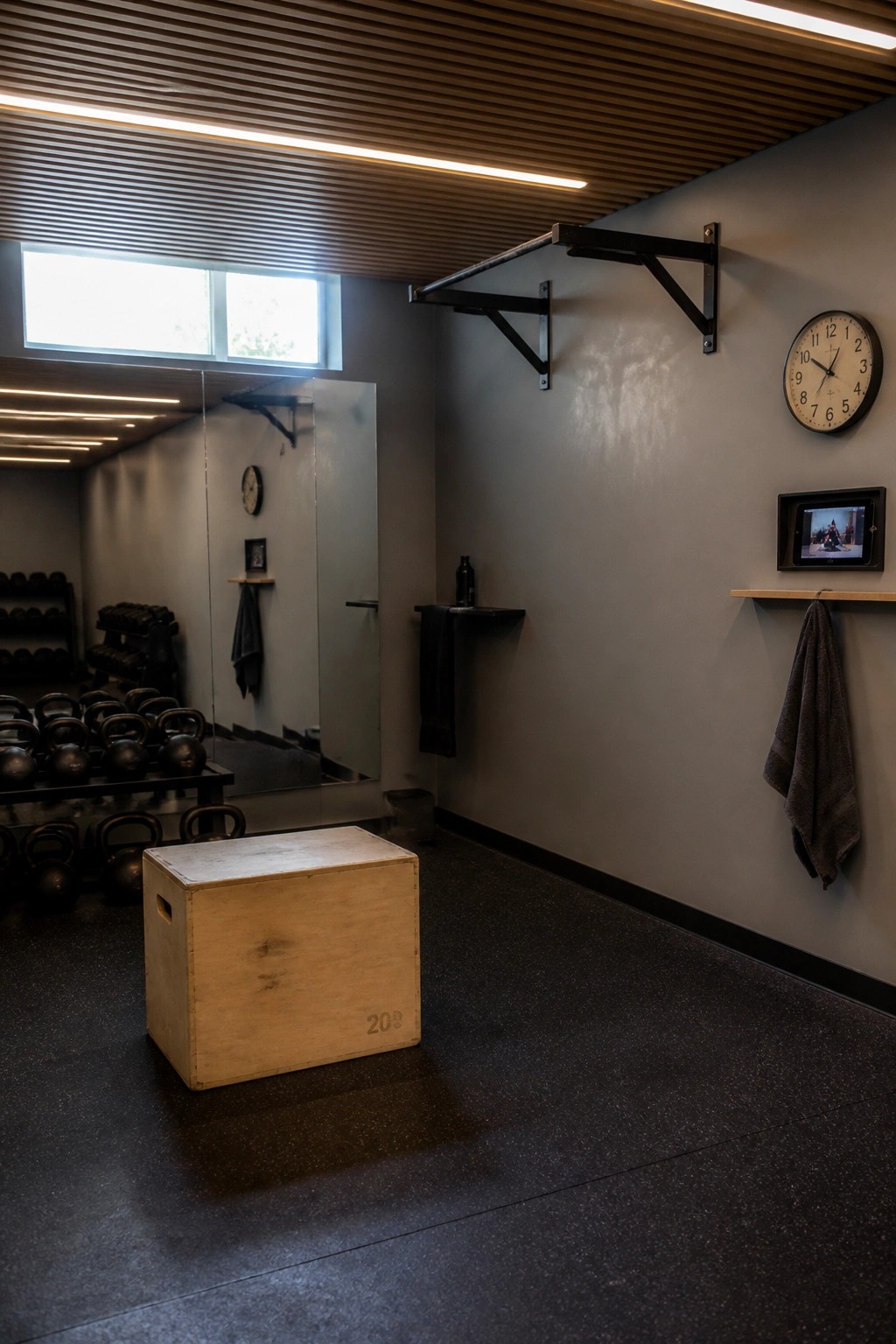Gym with wooden plyo box, pull-up bar, wall clock, and kettlebell racks reflected in mirror.