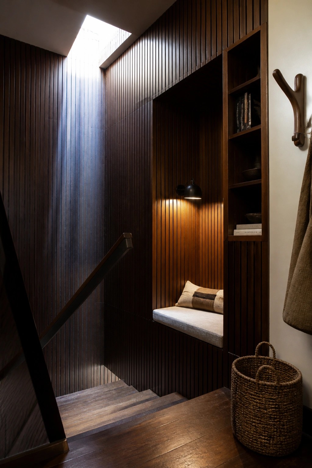 A narrow interior stairwell with dark vertical wood paneling includes a recessed cushioned bench, built-in shelves, and a skylight above the stairs.