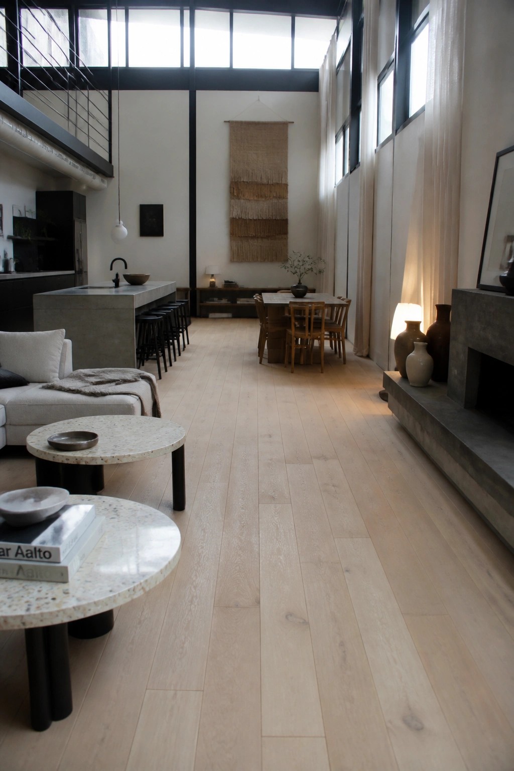 A wide open interior with light wood flooring running continuously through living, dining, and kitchen areas under high ceilings and large windows.