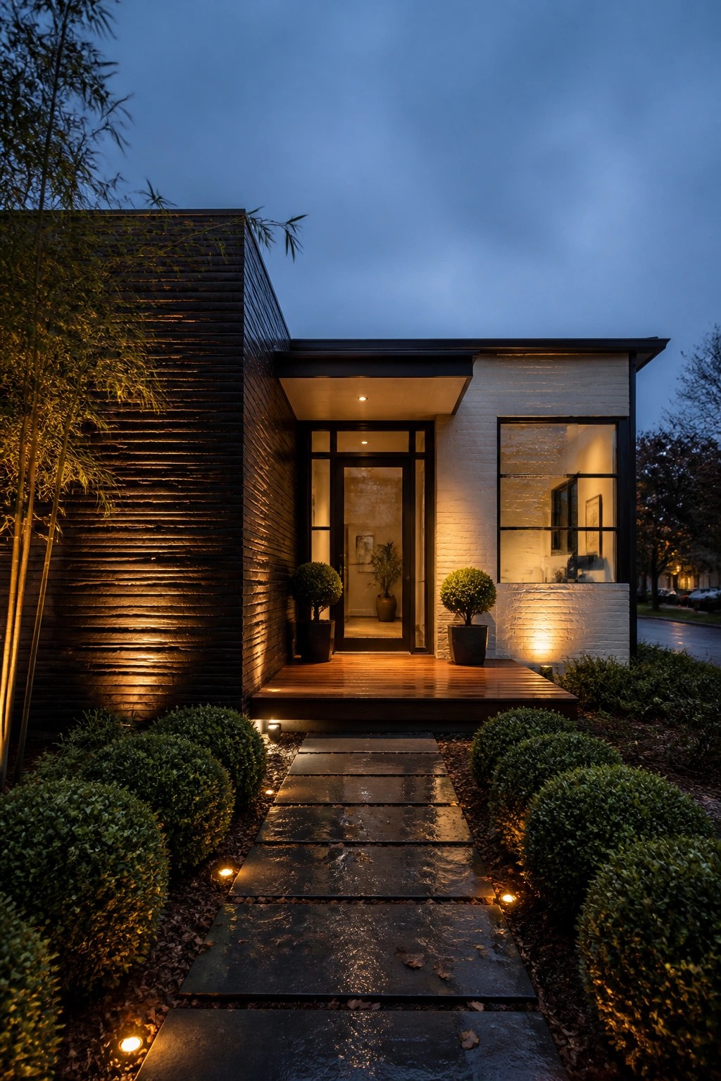 A modern house at dusk shows dark horizontal wood cladding on the left side of the facade and white brick on the right, with a lit entry door and a stone pathway in front.