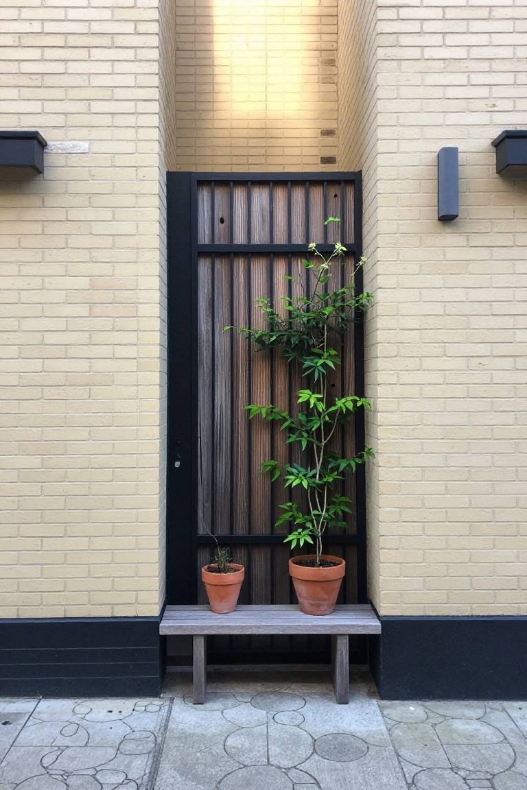 A black-framed door with vertical wood slats centered in a light brick wall, with two terracotta pots on a low bench below.