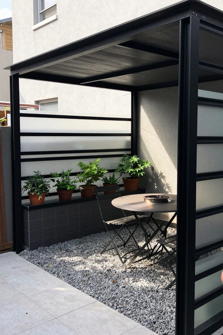 Outdoor seating area under a black metal structure with horizontal frosted panels, potted plants along a tiled base, and a round table with folding chairs on gravel.