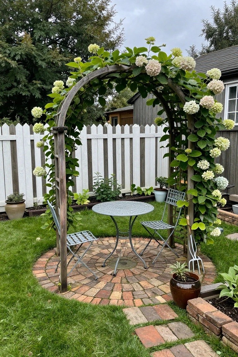 A white picket fence stands behind a vine-covered wooden arch over a round brick patio with a metal table and two folding chairs.