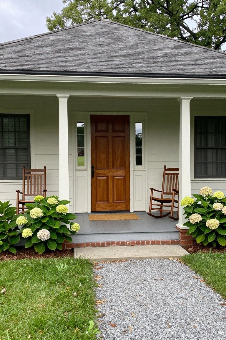 A white house with a brown paneled wooden front door centered on a porch between two white columns, with rocking chairs on each side and hydrangea bushes in front.