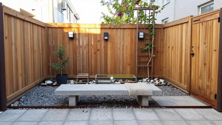 19 Decorative Wood Fence Ideas Featuring Planters, Panels, and Accents Modern wooden fence with black sconces, concrete bench, potted tree, and gravel bed.