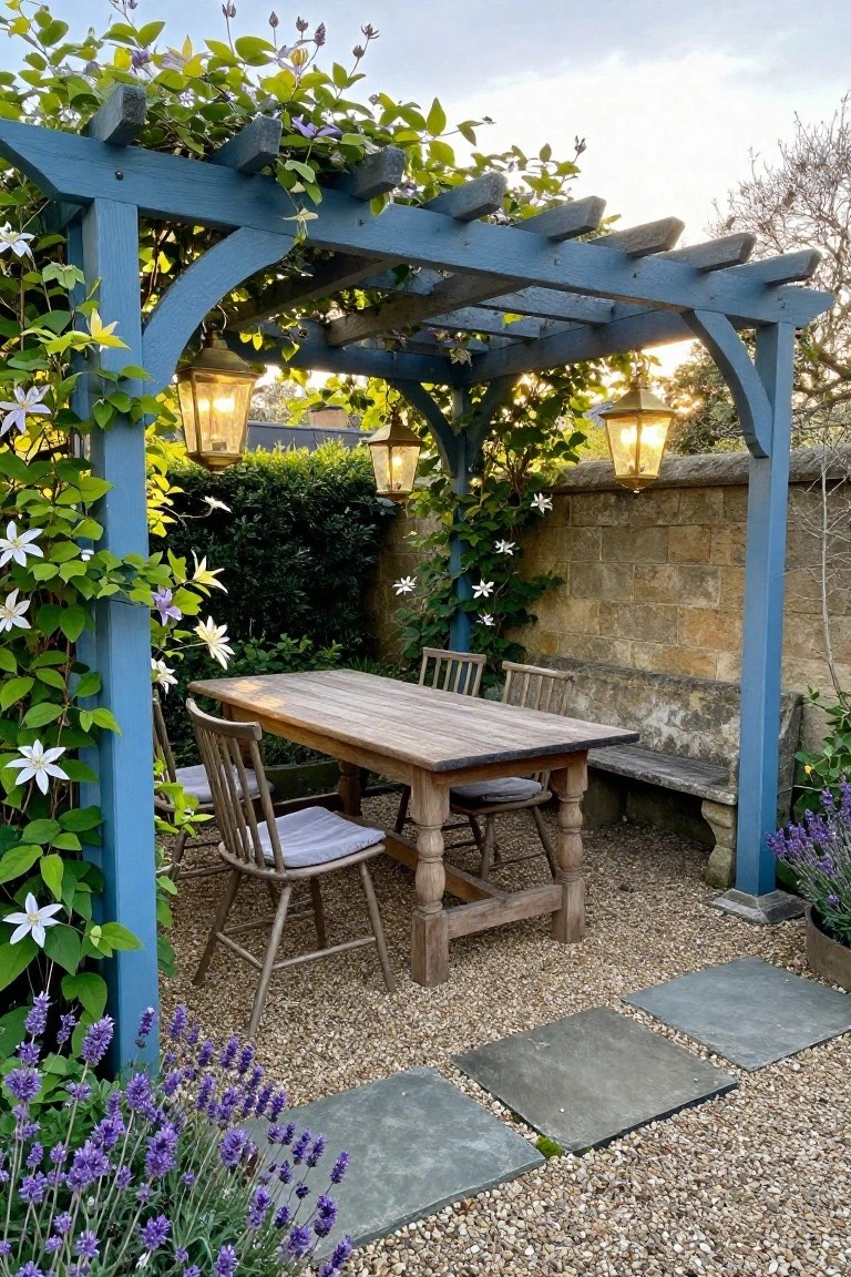 Blue wooden pergola sheltering wooden table and chairs with hanging lanterns amid flowers