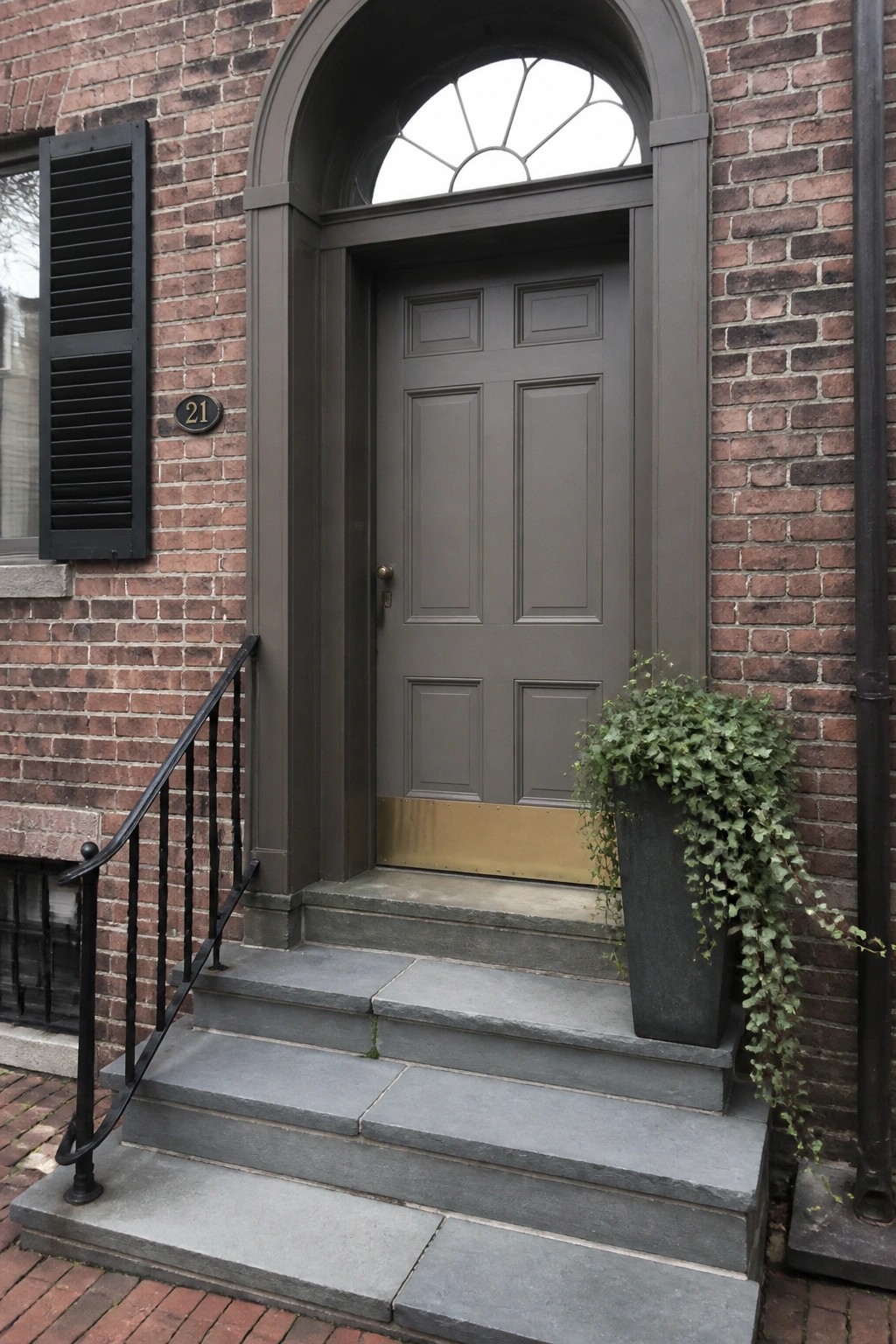 A gray paneled wooden door with a brass kickplate beneath an arched transom window on a brick house, with gray stone steps and a tall planter to the right.