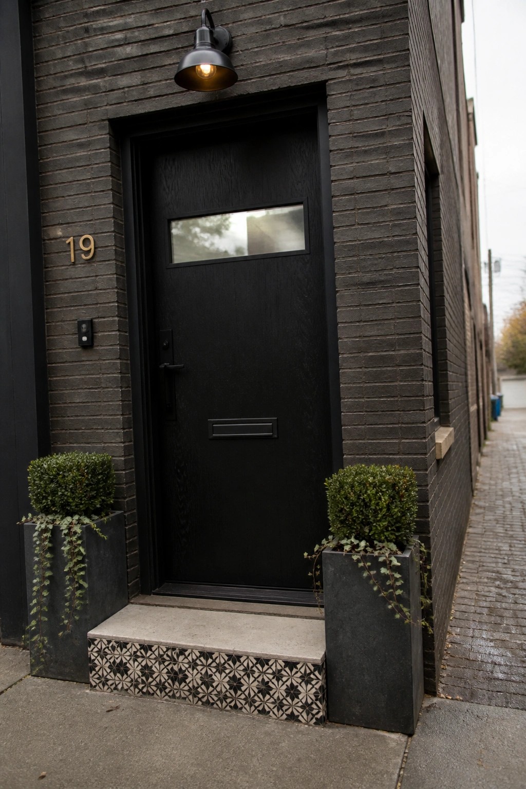 A black wooden door with a narrow transom window centered in a dark brick facade, with two concrete planters holding shrubs on either side of the step.