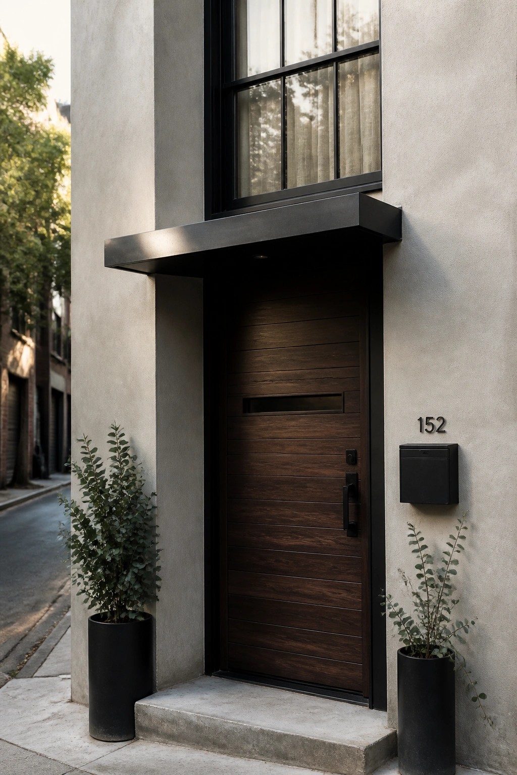 A modern house entrance with a dark wooden door featuring horizontal slats, a black metal overhang above, and two black planters with greenery on either side of the steps.