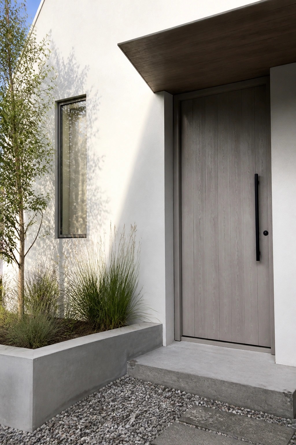 A modern gray wood door with a long black handle on a white house exterior next to a narrow window and a concrete planter with tall grasses.