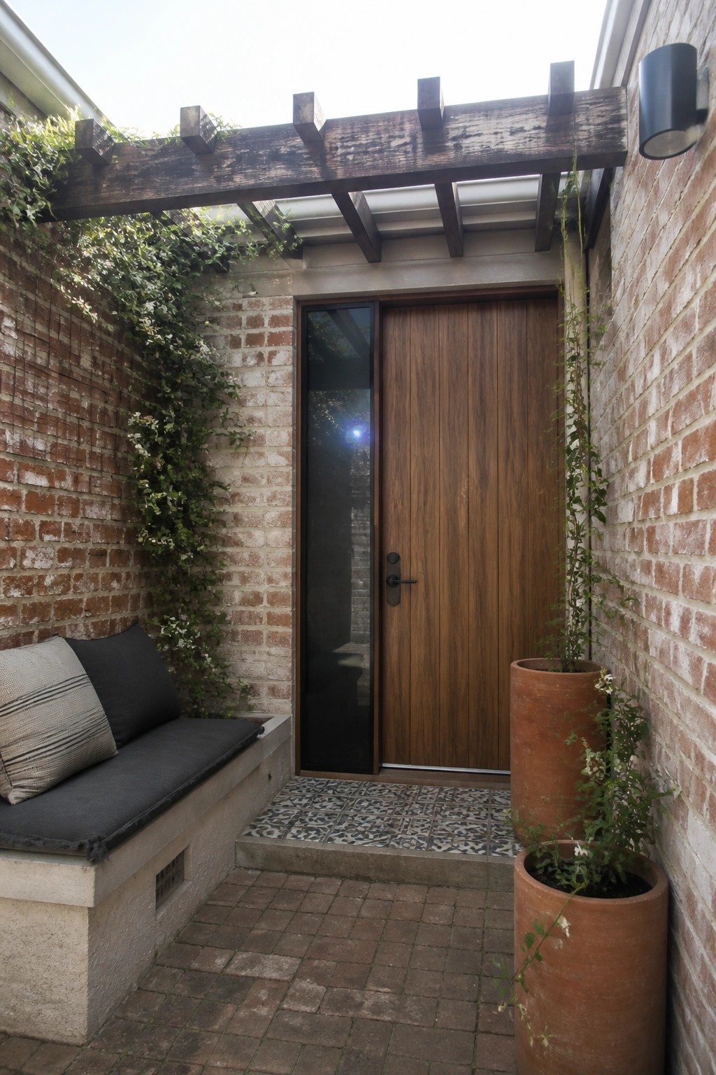 A vertical wood door with a narrow glass sidelight on the left, set in a brick-walled outdoor entry under a wooden pergola with potted plants nearby.