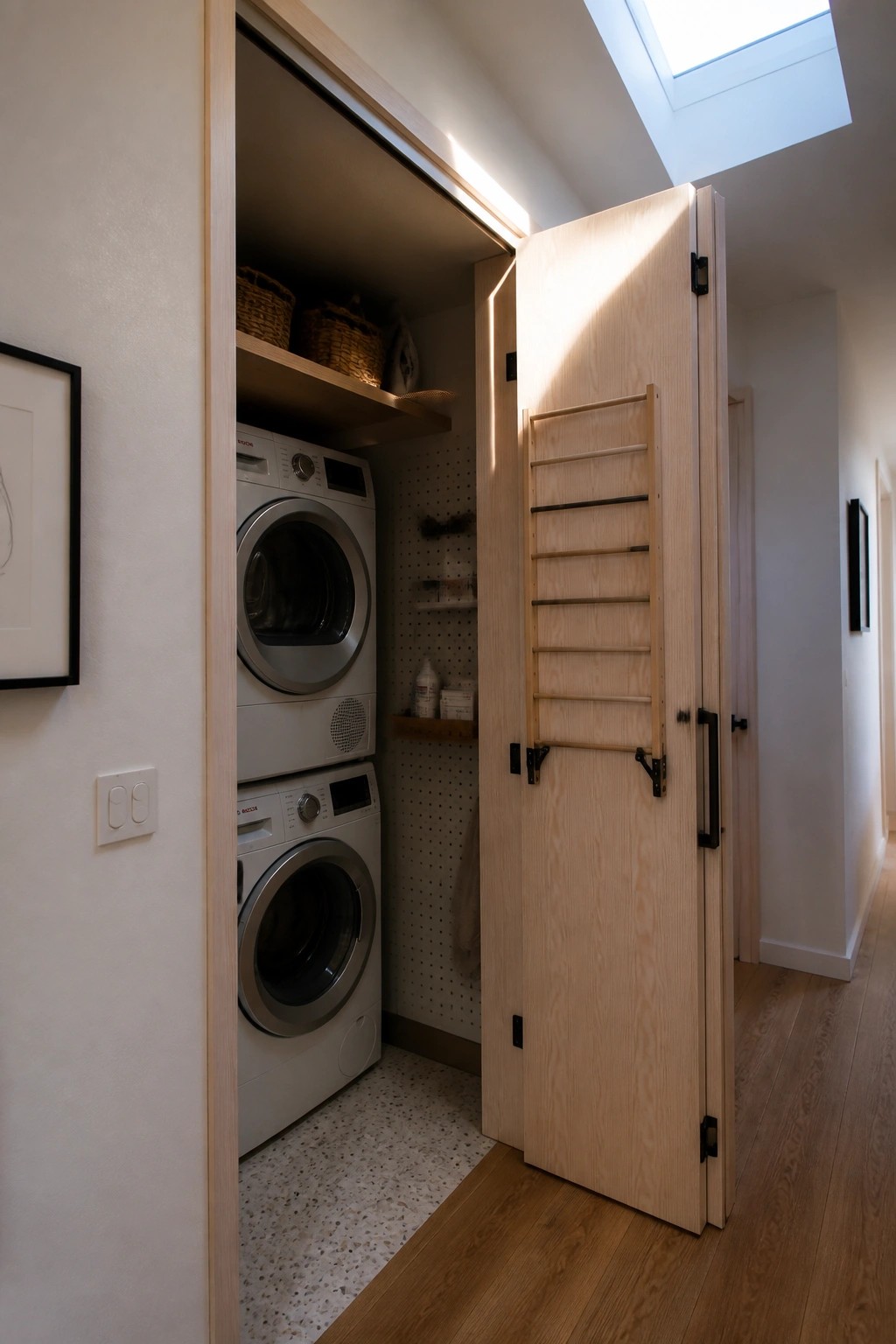 Light wooden folding doors stand open to reveal a stacked washer and dryer inside a narrow laundry space with a pegboard wall and wood floor.
