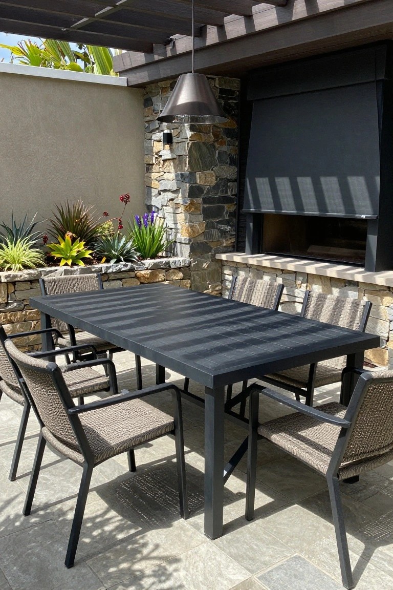 A dark rectangular patio table with woven chairs sits under a wooden pergola next to a stone outdoor fireplace.