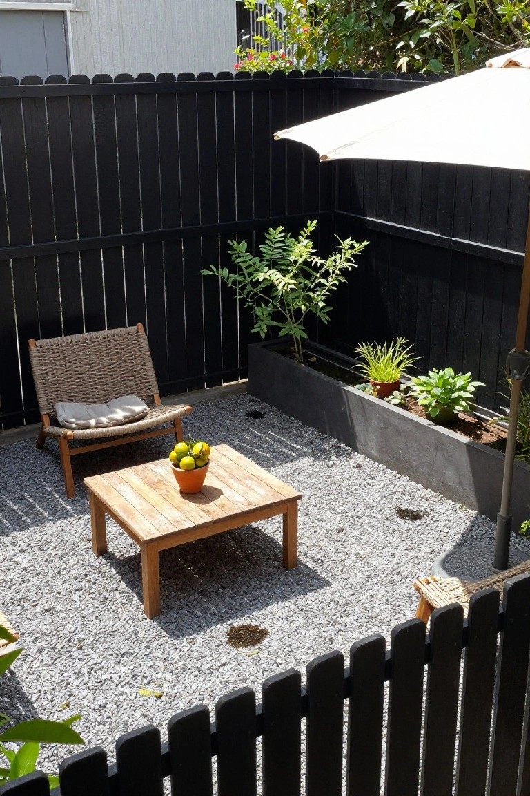 A black wood fence encloses a gravel patio with a woven chair, wooden table, and raised planter box holding small plants.