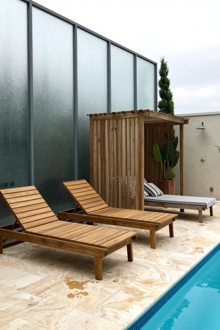 Two wooden lounge chairs sit in front of a tall vertical wood slat screen beside a swimming pool on a stone patio.
