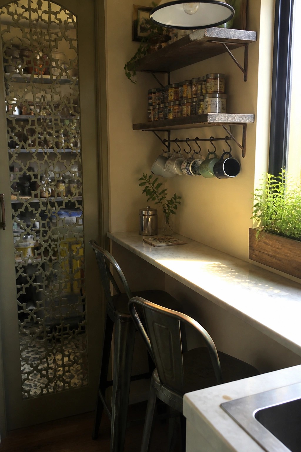 A carved wooden jali door stands beside a kitchen counter with two metal stools, open shelves holding spice jars, and hanging mugs near a window.