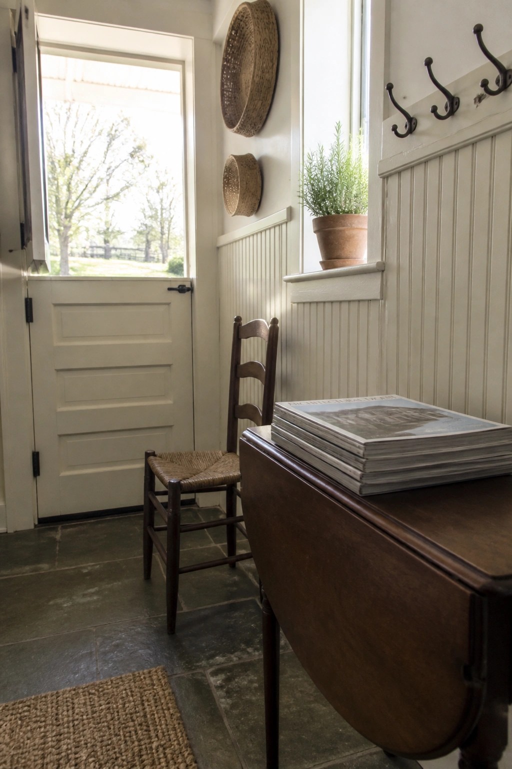 An open white paneled wooden door stands beside a dark wood chair with a woven seat in a room with stone floors, wall hooks, and a potted plant on the windowsill.