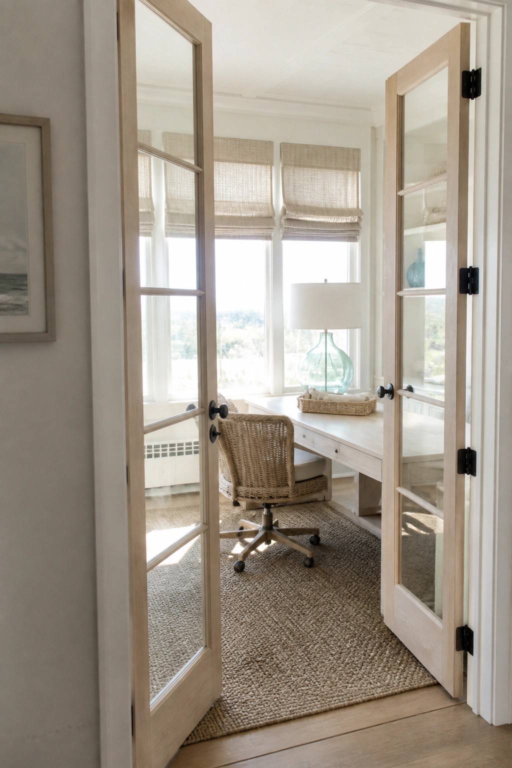 Open light wood French doors with glass panels leading into a home office with a white desk, woven chair, and windows with woven shades.