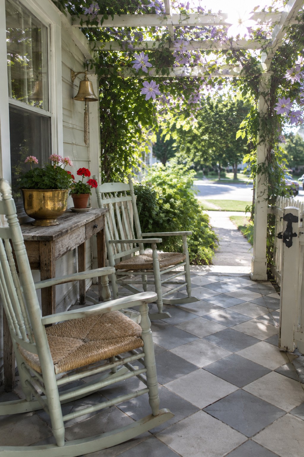 A white wooden pergola covered in green vines and purple flowers shades two light green rocking chairs on a checkered tile porch beside a house.