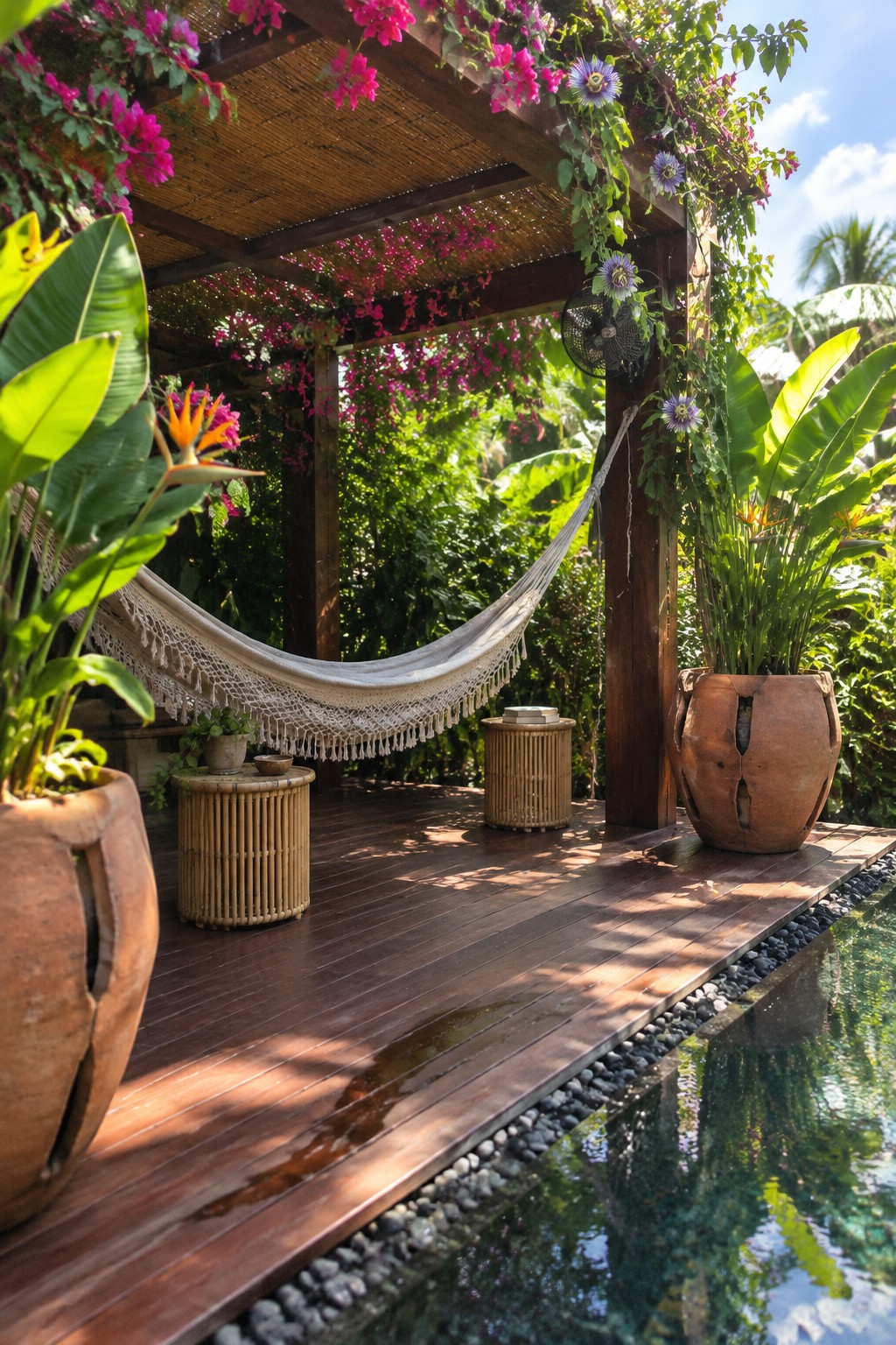 A wooden pergola covered in pink and purple flowering vines stands on a dark deck next to a pool, with a white hammock hanging underneath and large potted plants nearby.