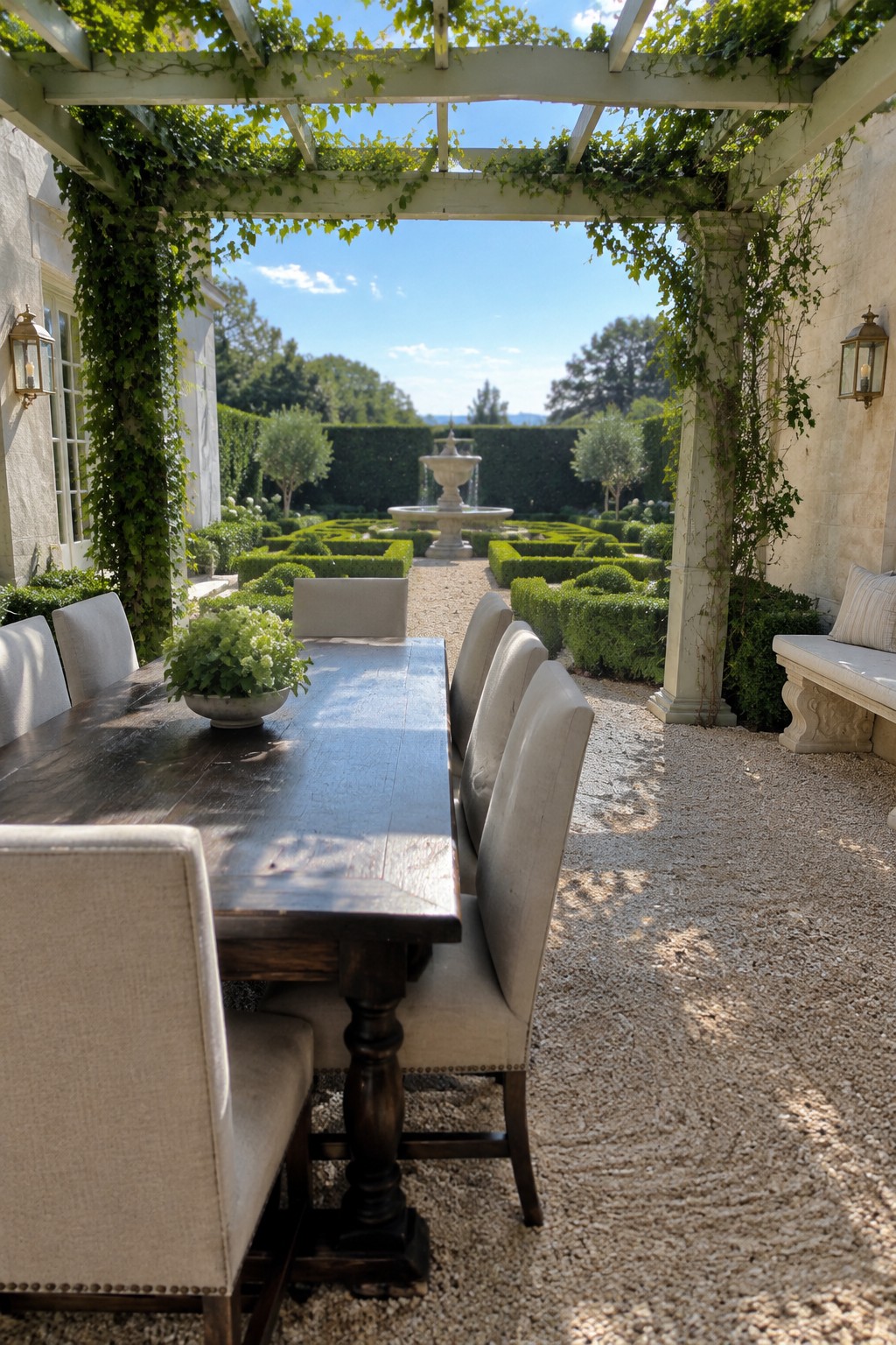 A wooden pergola draped in green vines covers a long dining table and chairs on gravel, with a formal garden and fountain visible in the background.