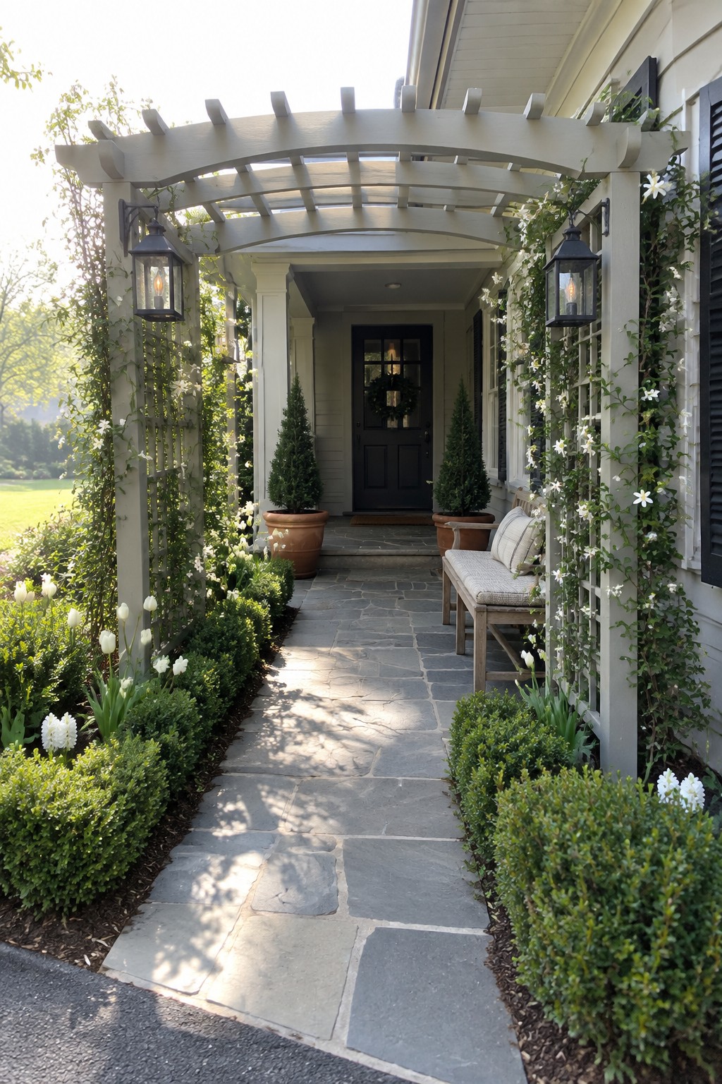 Stone pathway under vine-covered white pergola leads to black door and bench