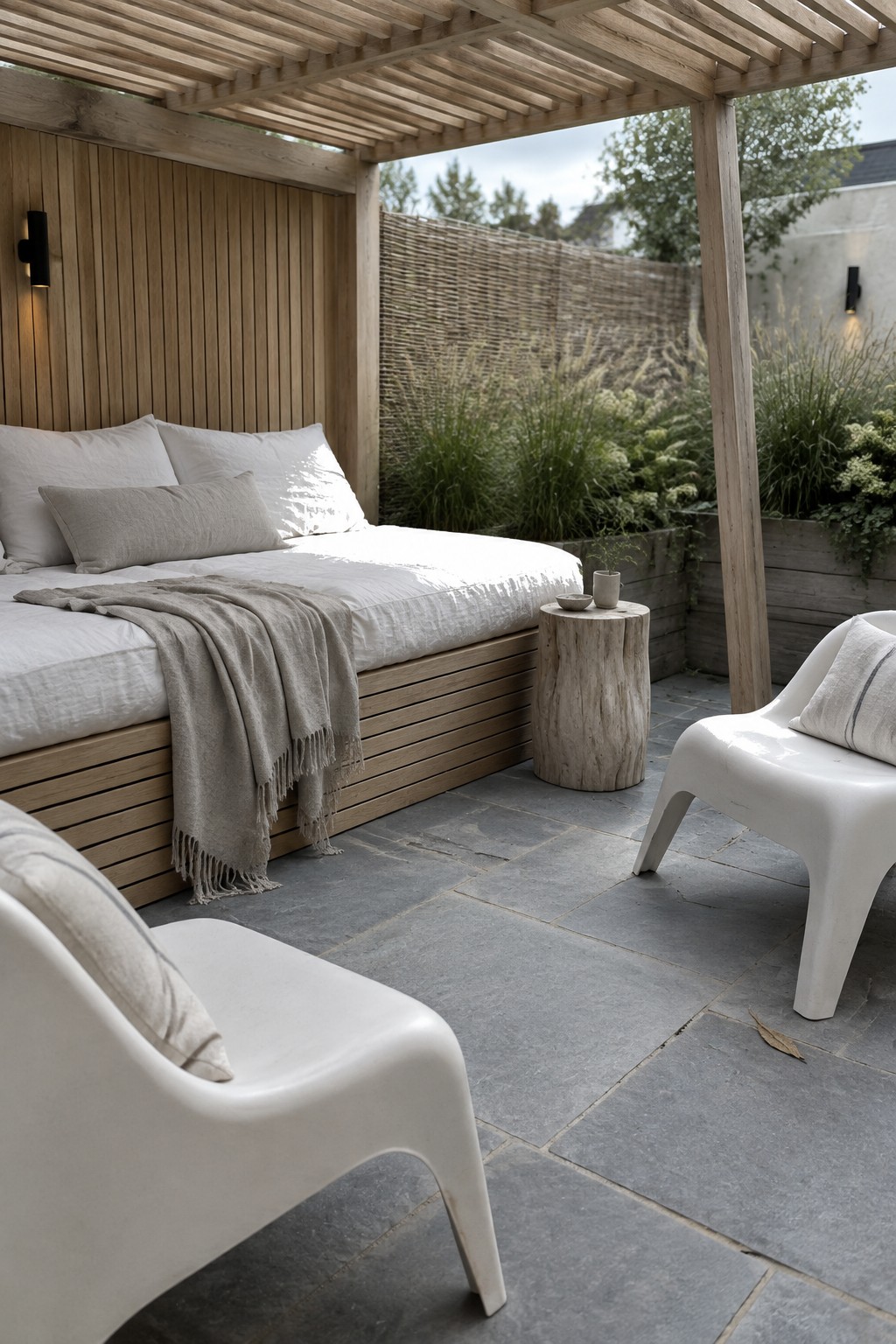 A wooden pergola shades a built-in daybed with white linens and pillows, set on a stone patio beside two white outdoor chairs.