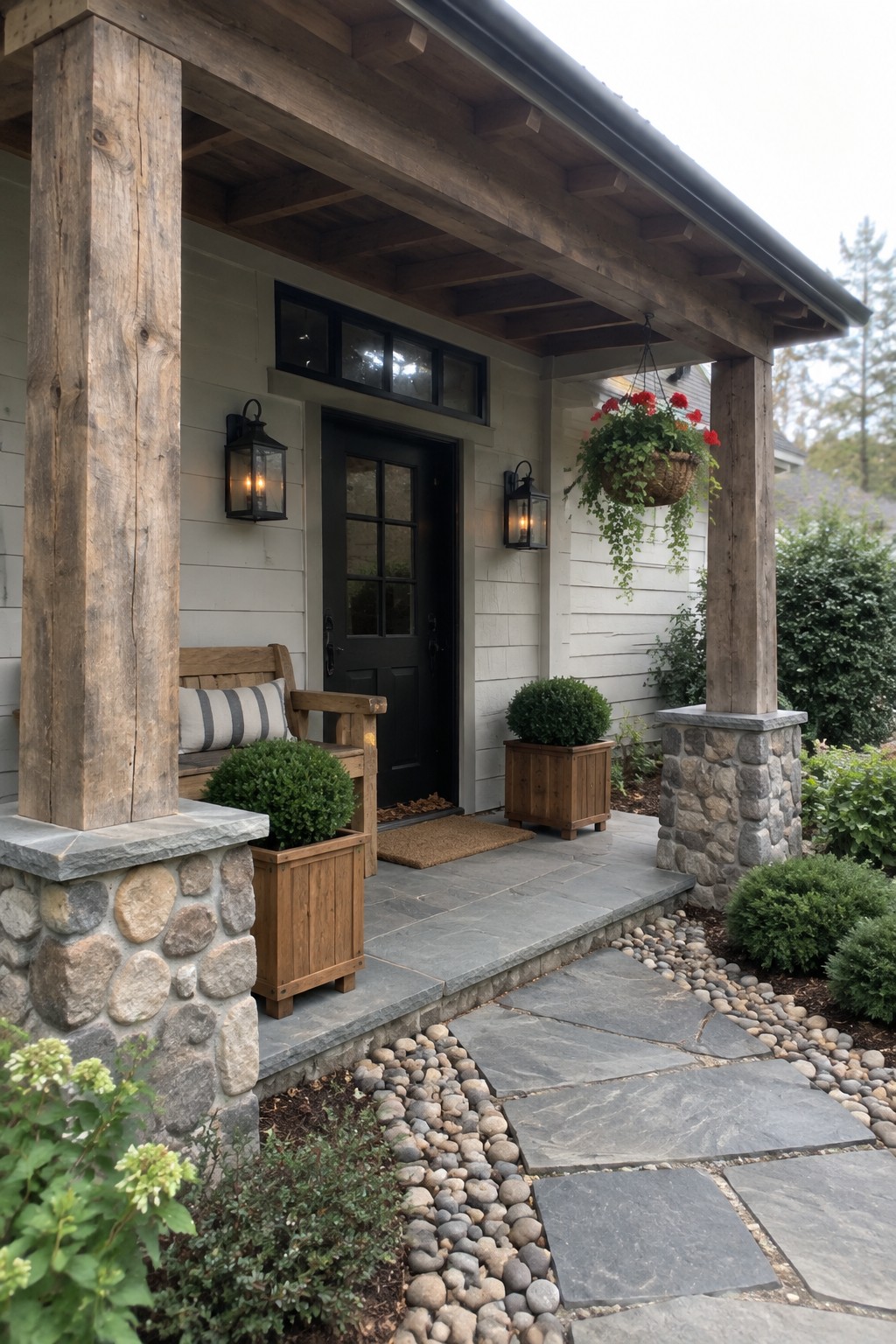 A house entry porch with thick wooden beams supported by stone pillars, a black door, and a stone pathway bordered by pebbles.