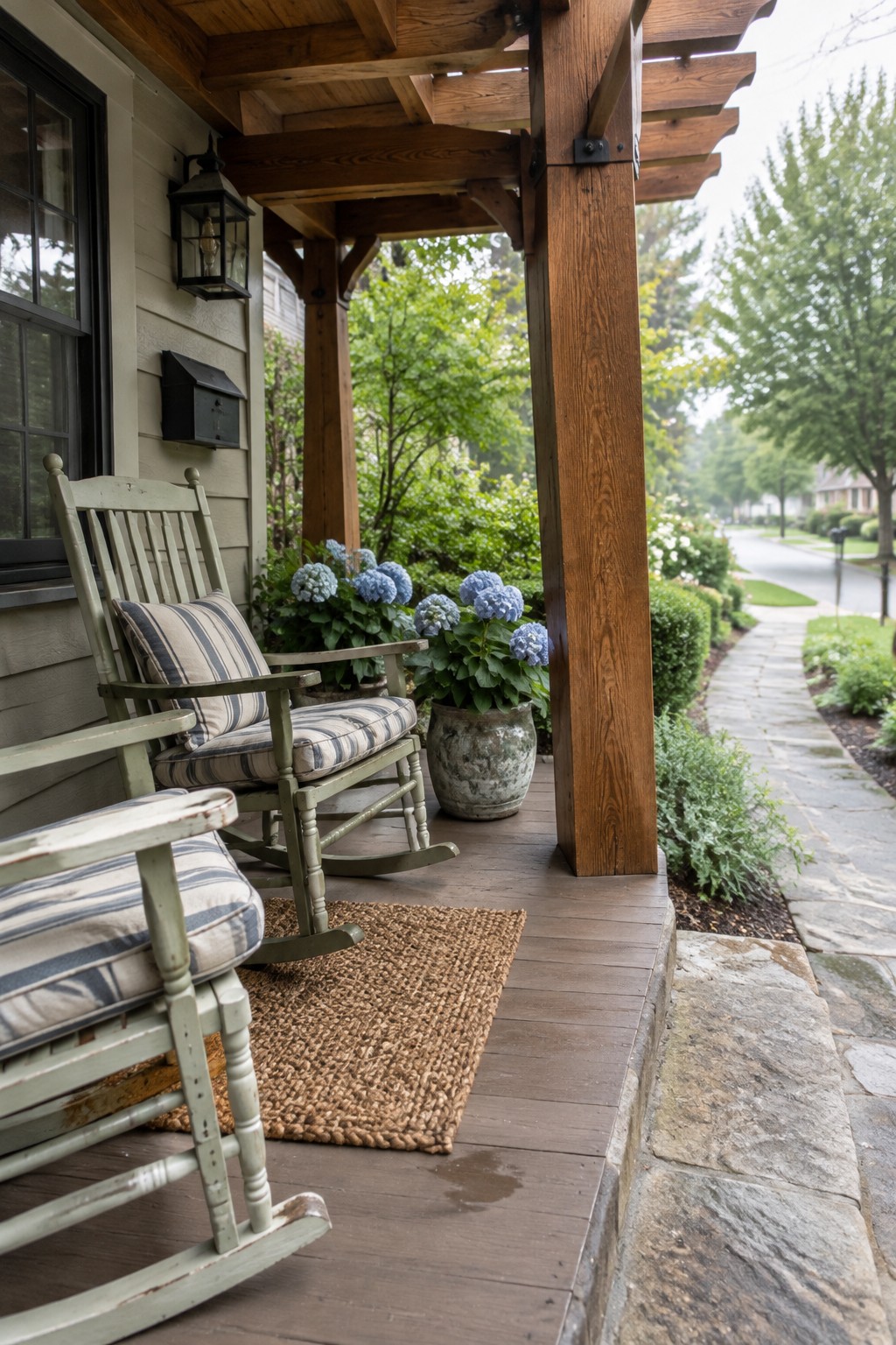 A wooden cedar pergola covers a front porch with two green rocking chairs, striped cushions, and potted blue hydrangeas beside a stone path.