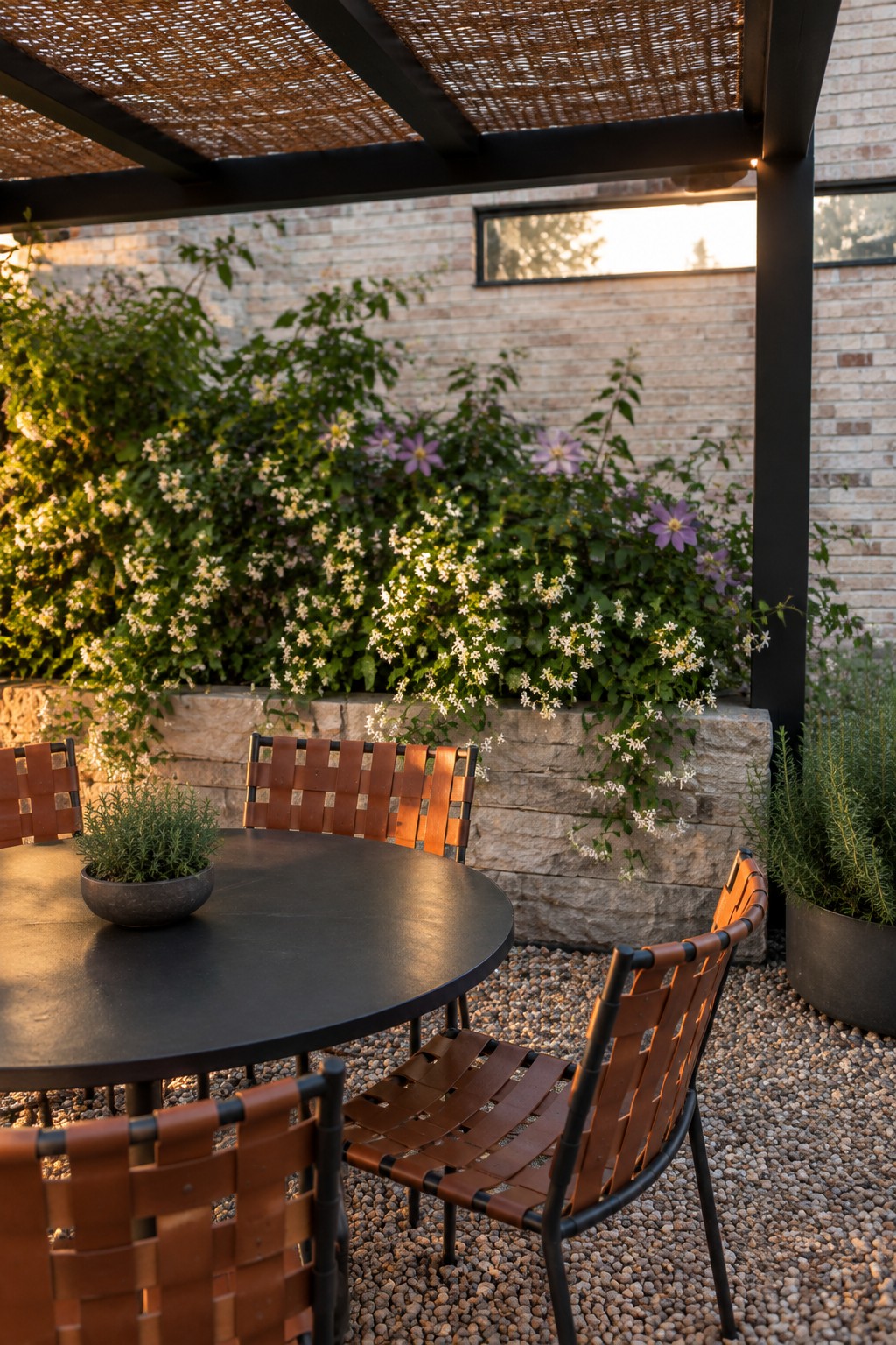 A black metal pergola with a woven brown shade covers a round black table and brown leather strap chairs on gravel, beside a low stone wall with white and purple flowers and a brick house in the background.