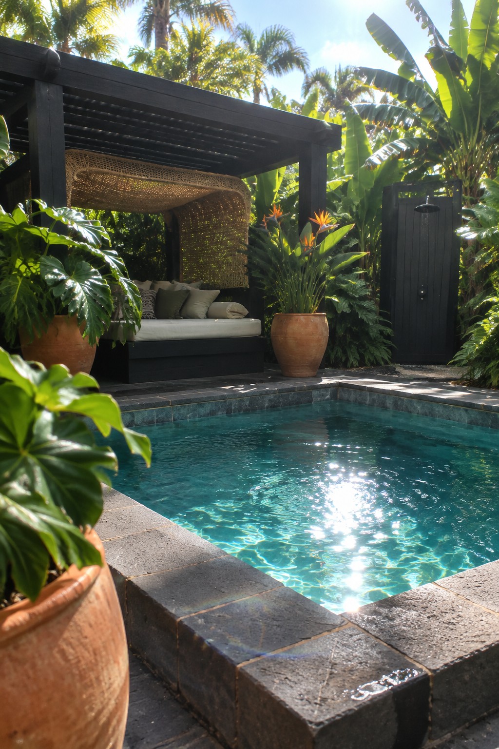 A black wooden pergola with a daybed beneath it sits beside a swimming pool in a tropical garden setting.