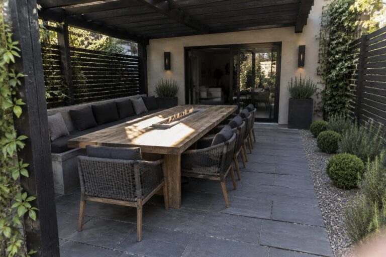 Modern patio with long wooden table, lit fire pit, woven chairs under dark pergola