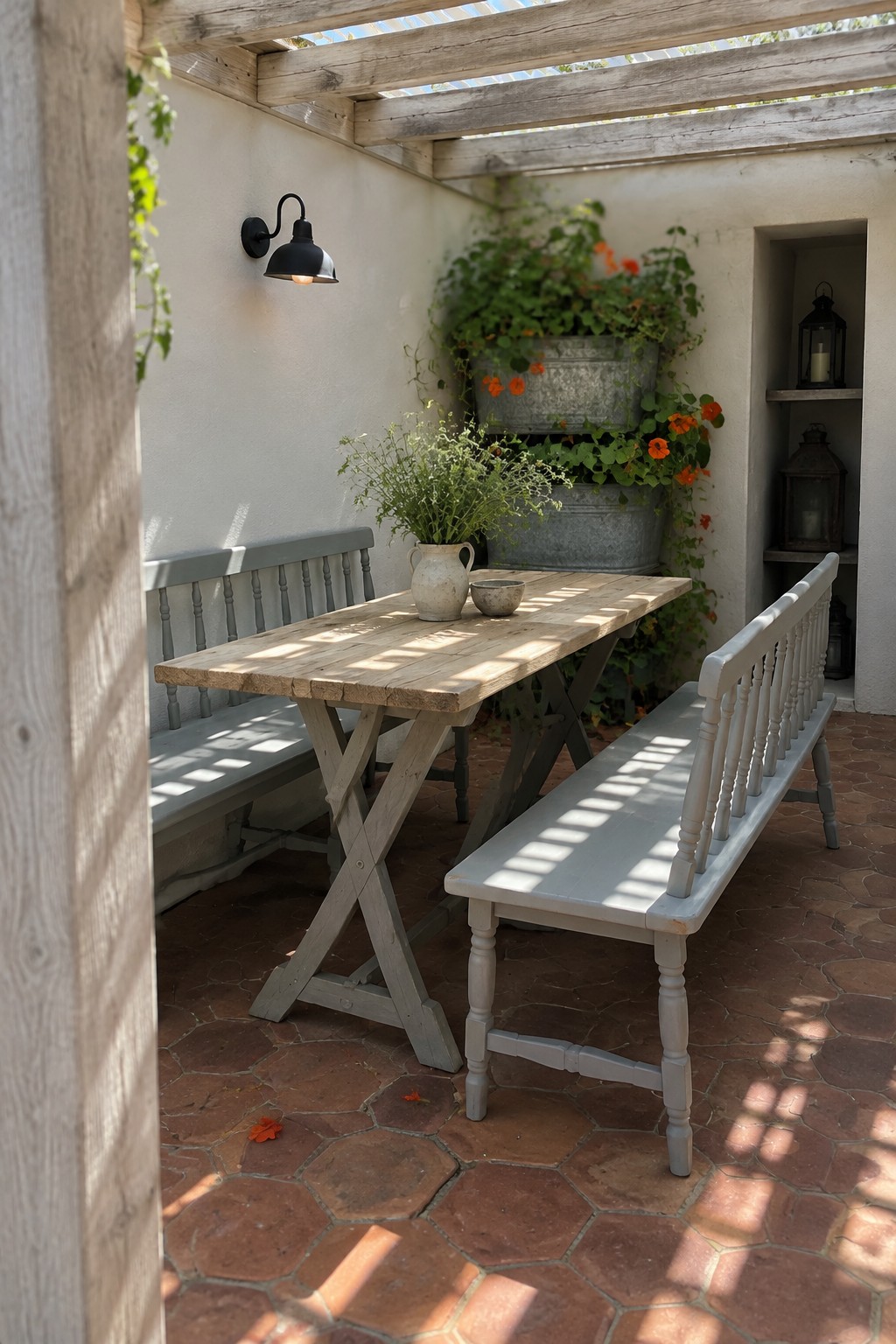 A small wooden pergola with open slats covers a compact brick patio furnished with two light gray benches and a wooden table holding pottery, surrounded by potted plants and climbing vines.