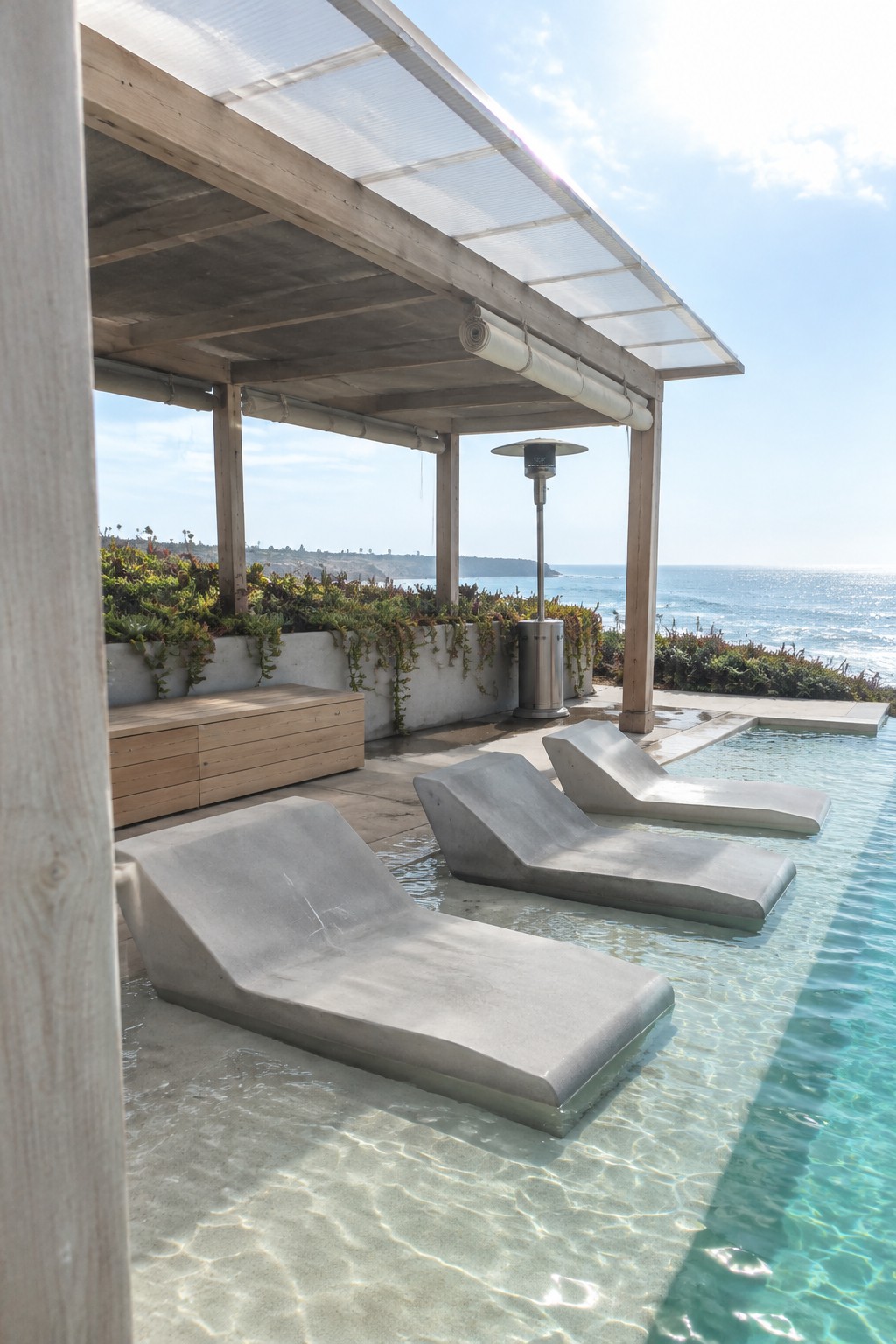 Concrete lounge chairs sit partially in shallow pool water under a wooden pergola with a translucent roof, overlooking the ocean.
